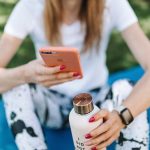 Woman using smartphone while holding bottle on yoga mat outdoors.